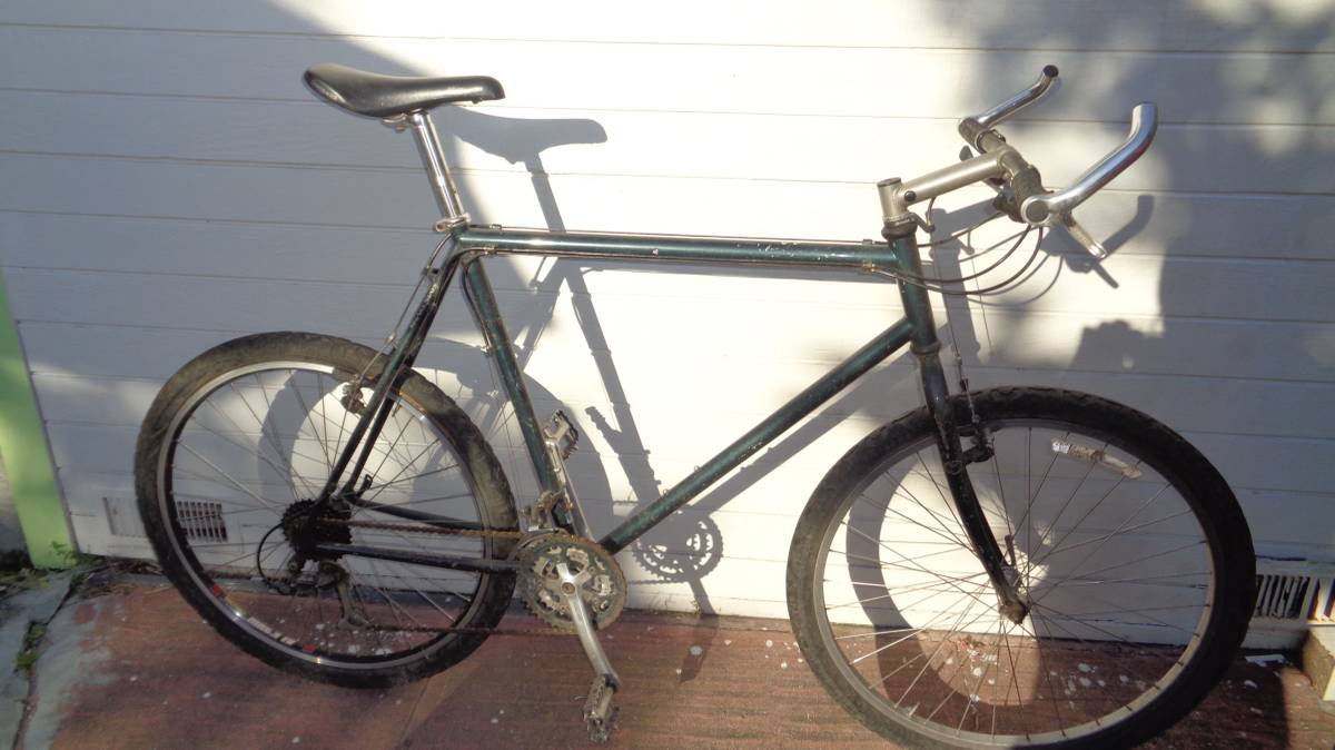 a large 1994 Diamond Back mountain bike with evergreen job stands on a porch. The bike has flat bars, knobby 26" tires, and is a little dirty.