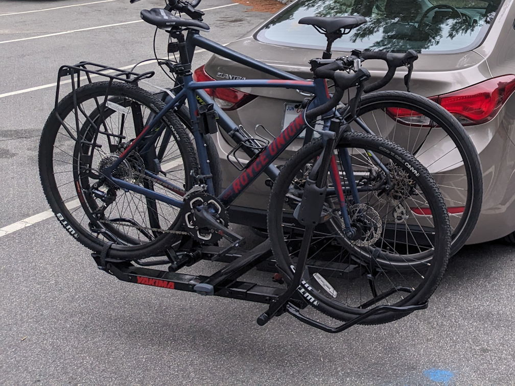 A blue gravel bike on the rear hitch rack of a tan Hyundai Elantra, with a Specialized hybrid bike behind it