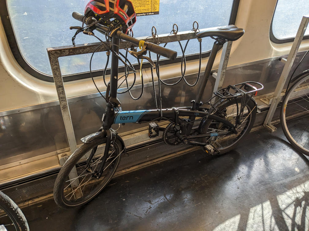 A Tern Link D8 folding bike in black and blue livery is on a train bike car, being held in place to a rack with a rubber bungee. A red Specialized helmet sits on the handlebars.