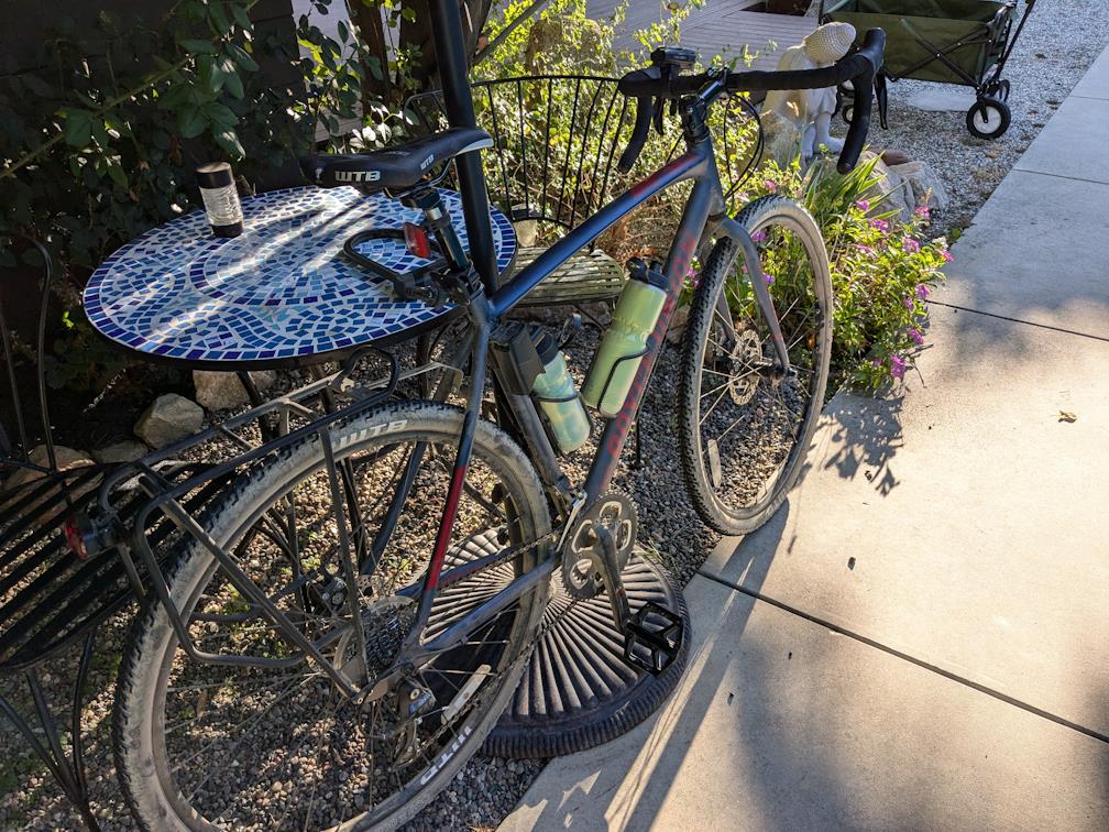 a bike with drop handlebars, knobby tires, two waterbottles in holders, and a rear rack sits next to an outdoor bistro table. Shadows are long in the morning light.