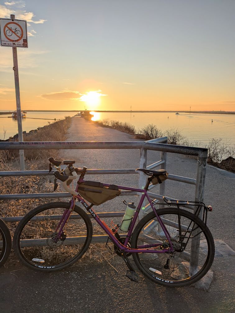 a bike leans against a fence along a jetty with the sunset behind. The bike is purple with orange accents and has drop handlebars, knobby tires, various frame bags, and a rear rack