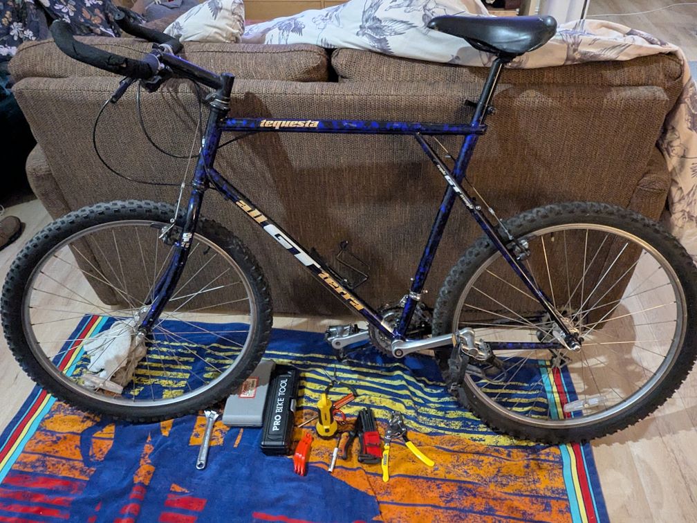 a large 1994 GT Tequesta mountain bike with blue/black paint job leans against the back of a sofa. The bike has bull bars, knobby 26" tires, and is in ok shape given it's 30 years old.