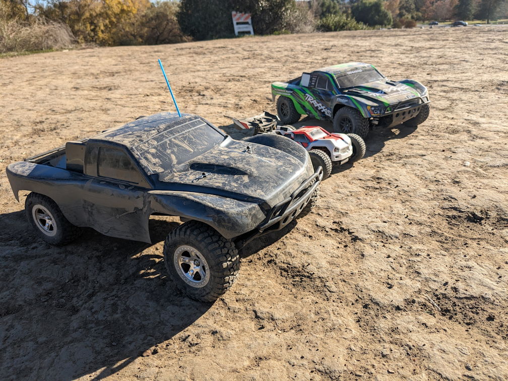 three dusty RC cars (a black 2wd slash, a red reflex 14t, and a green slash 4x4) sit posed next to each other in a dirt clearing
