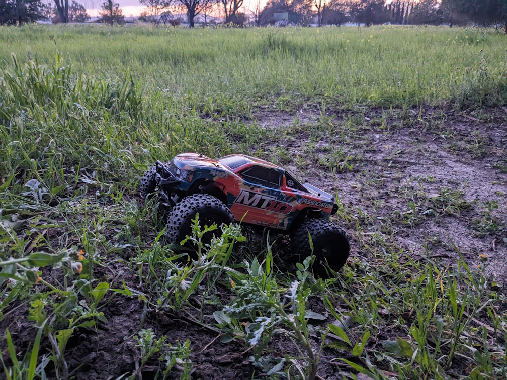 a photo of a blue and orange camo Rival MT10 monster truck facing upward on an overgrown dirt ramp