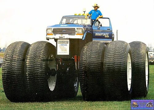 the biggest version of the bigfoot monster truck ever created, with dual wheels from the Army's failed Land Train project. a main with a cowboy hat and sunglasses leans out the open door from the driver's seat