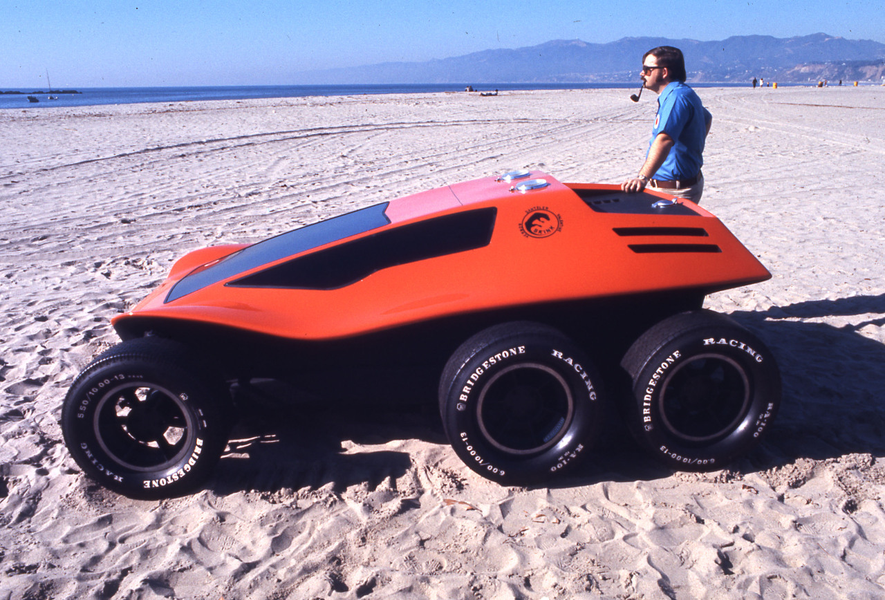 an experimental 6-wheeled beach buggy sits on santa monica beach, a man sits astride