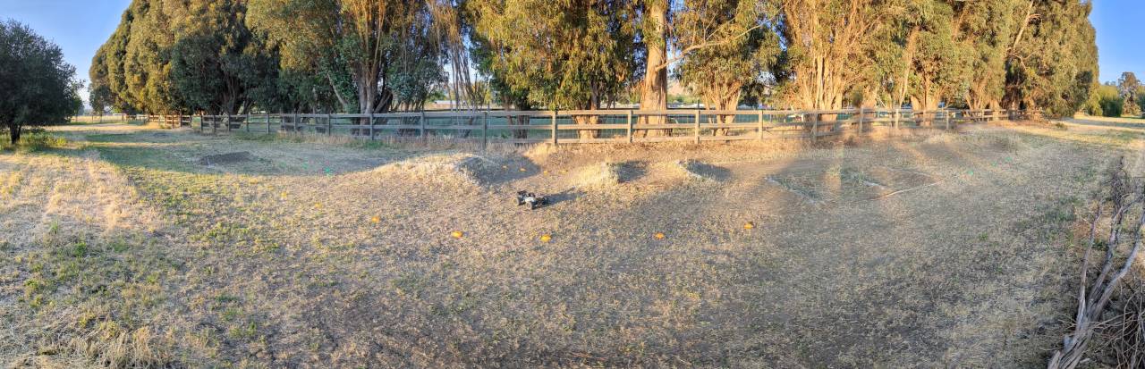 a panoramic shot of a backyard rc track with a tekno mt410 monster truck parked on it, dirt and scrappy weeds and grasses and an array of jumps and turns make up the rough motocross style course 