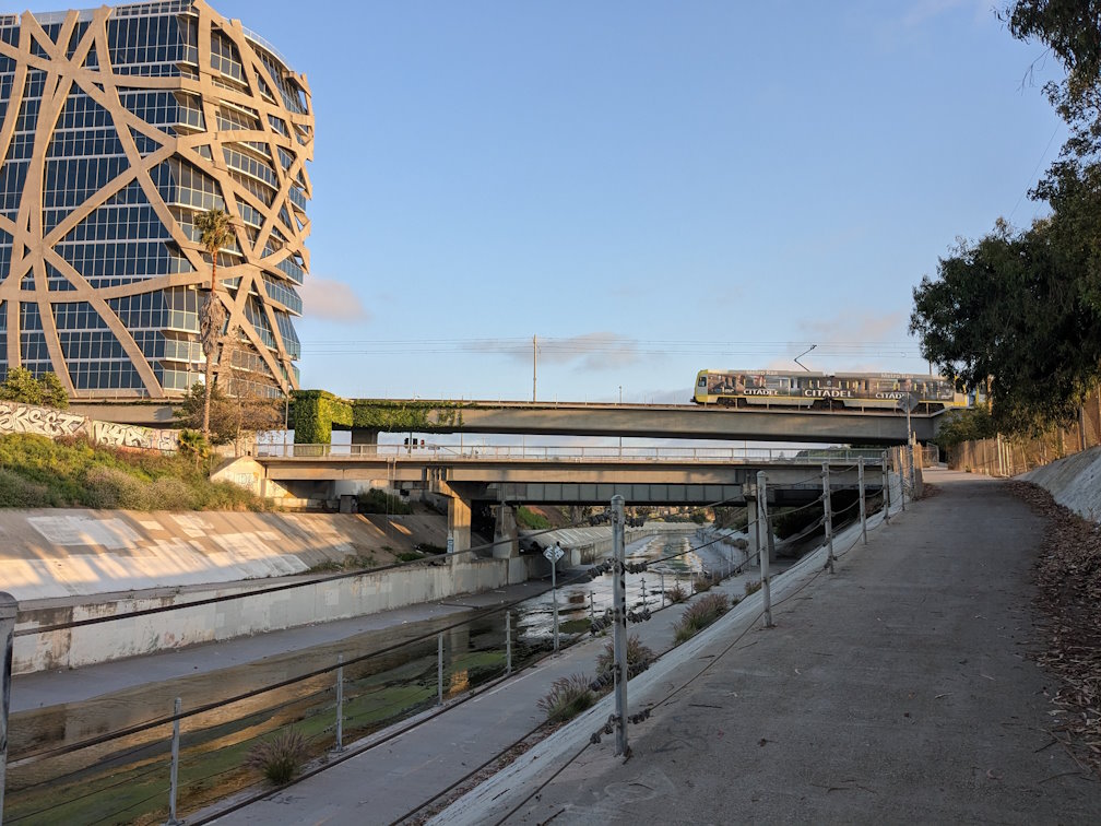 A concrete creek channel with a light rail train passing over it and the Wrapper building standing next to it